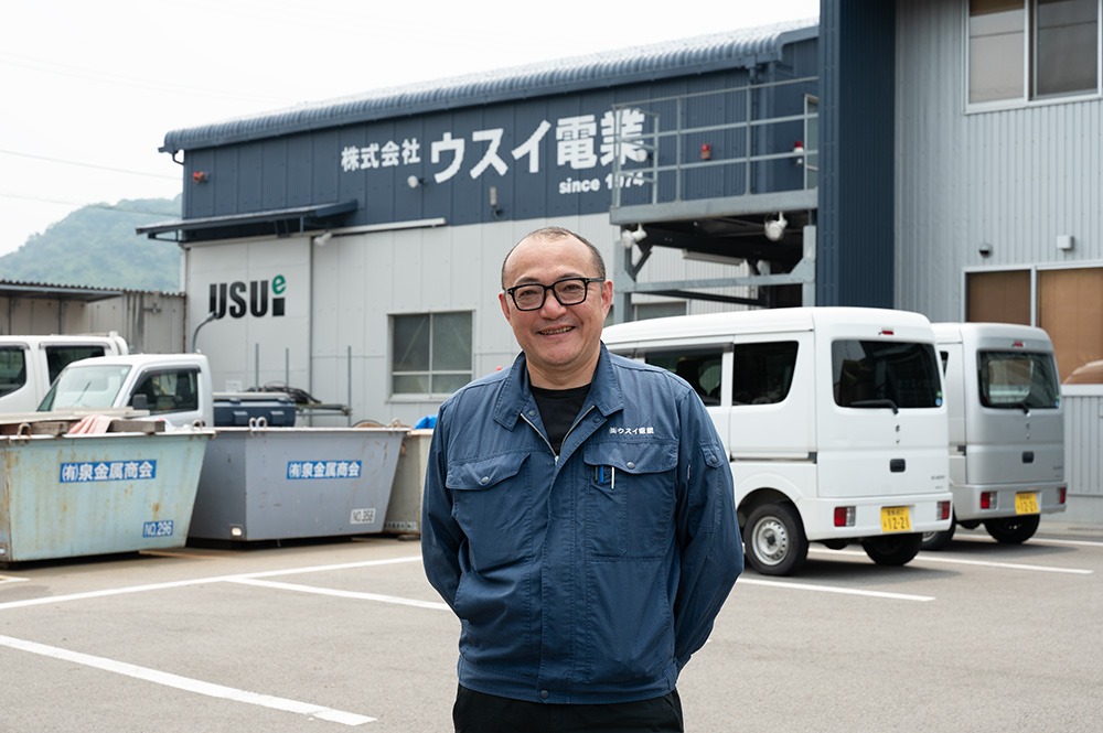 工場を背景に青い作業服を着た男性が立っている画像